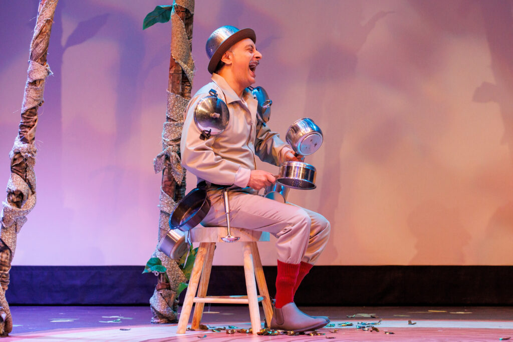 an actor sitting on a chair laughing, while many cooking-pots are hanging from his suit.