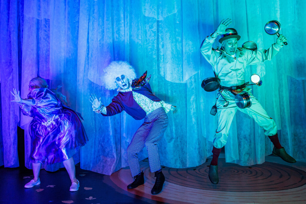 three actors standing on a stage that is covered in blue light, while seemingly being frightened.
